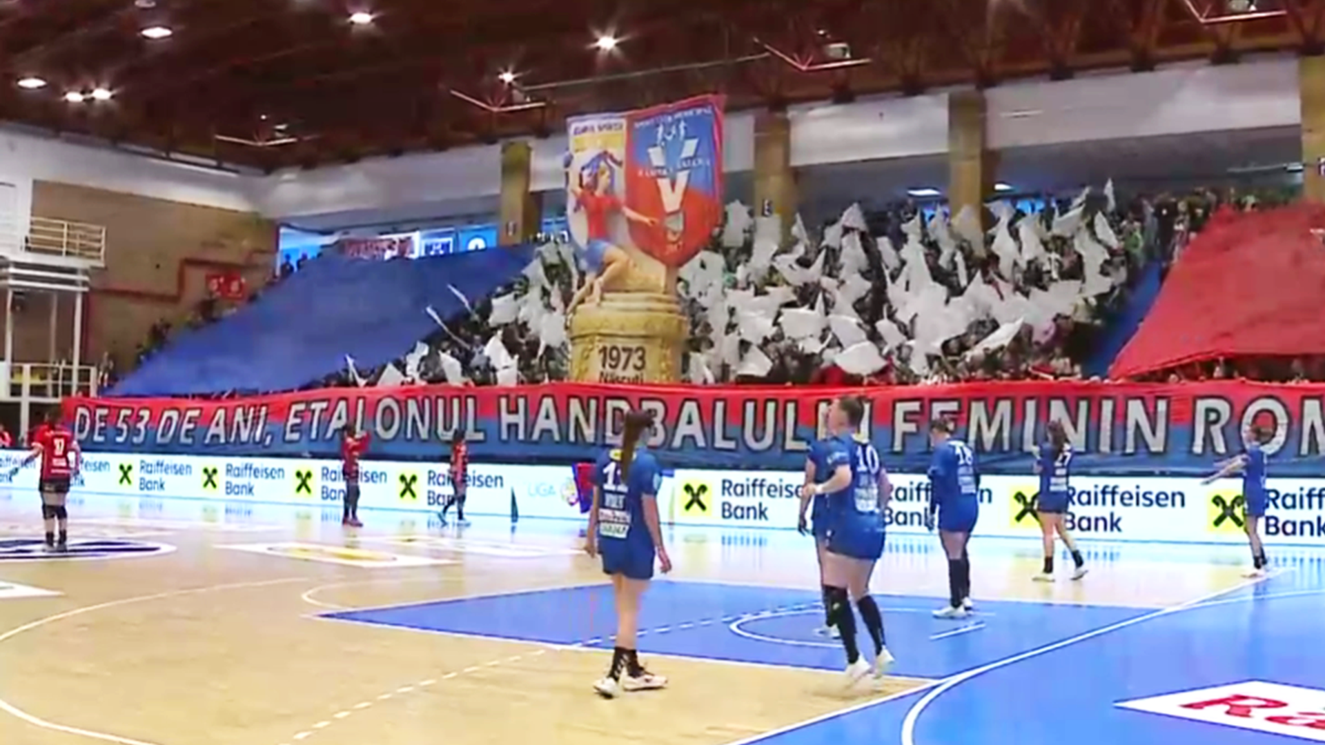 Jucătoare de handbal feminin de la Gloria Bistrița și SCM Râmnicu Vâlcea în acțiune în timpul meciului decisiv pentru titlu, cu tabela de scor vizibilă și galerie în fundal. | sursa foto: Gazeta Sporturilor